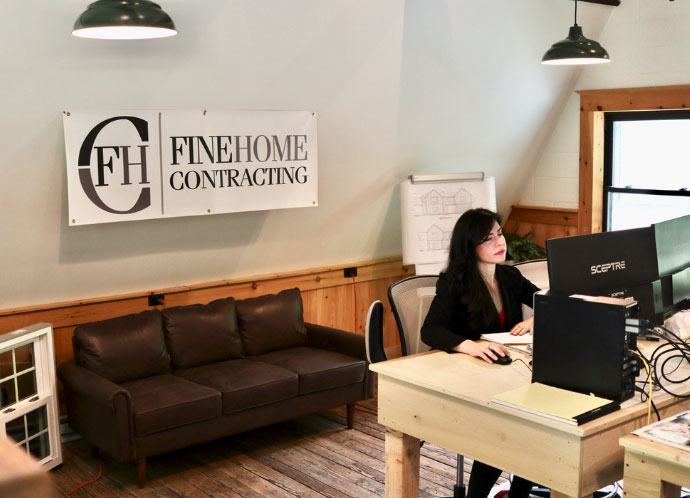 A woman works at a desk in an office with a "Fine Home Contracting" sign on the wall, a couch, a table, and architectural drawings.