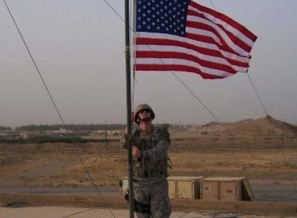 A soldier in uniform raises the American flag on a flagpole with a desert landscape in the background.