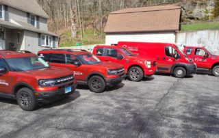 Four red service vehicles with company logos are parked in a row on an asphalt lot near a house and wooded area.