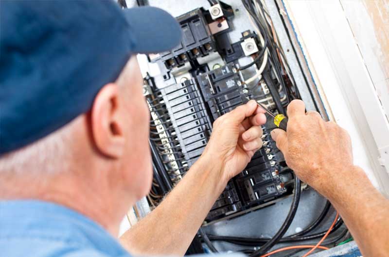 An electrician wearing a blue cap works on a circuit breaker panel, using a screwdriver to adjust wiring connections.