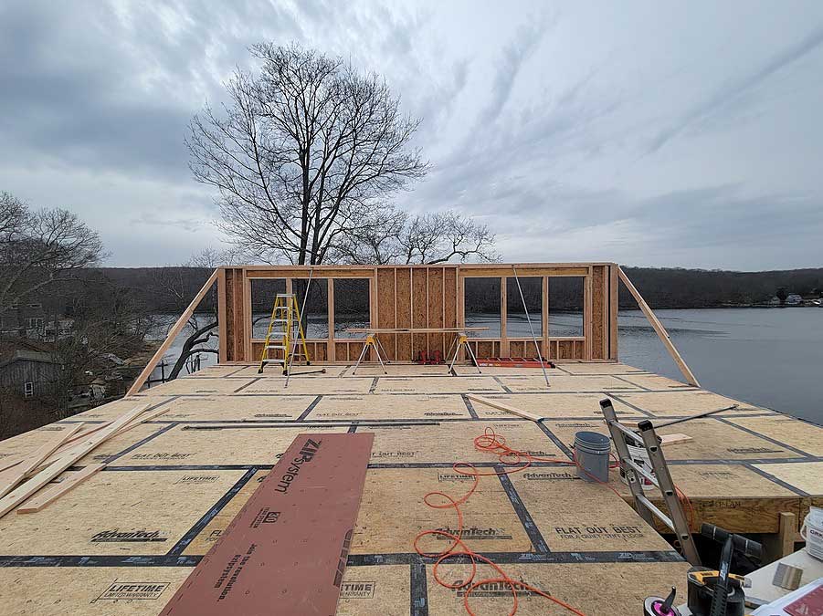 Partially constructed wooden frame of a second story addition with tools and a ladder on the floor, overlooking a body of water and trees.