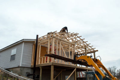 A person works on the wooden roof frame of an under-construction house extension, a testament to meticulous exterior remodels, with construction machinery visible nearby.