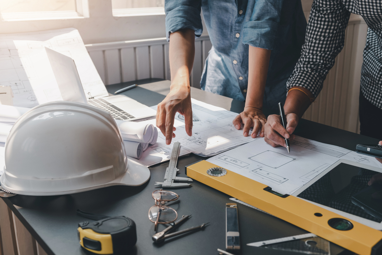 Two people working on architectural blueprints at a desk with a hard hat, caliper, glasses, tape measure, and level tool, showcasing the precision and collaboration of a design-build contractor.