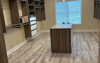 A spacious walk-in closet featuring wood shelving and drawers, an island in the center, and a large window with blinds.
