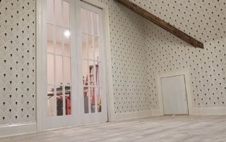 A room with patterned wallpaper, a wooden ceiling, a modern light fixture, and glass-paneled doors leading to a walk-in closet with wooden floors.