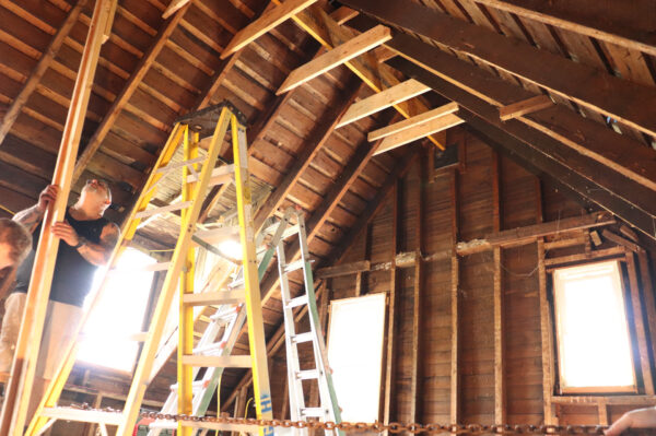IMG_1210 A person holding a wooden beam stands beside a ladder in a construction site with exposed wooden beams and an unfinished ceiling, referencing the Home Addition Planning Guide for detailed steps.