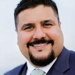 unnamed A man with short hair and a beard is smiling at the camera. He is wearing a suit and tie.