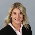 sarah A woman with long, blonde hair wearing a black blazer and a light pink blouse smiles against a plain grey background, radiating the confidence often seen in the best home inspectors.