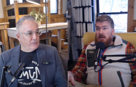 Two people are seated and speaking into microphones during a podcast recording in a room with shelves and various equipment.