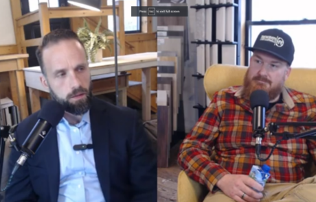 Two men sit in a studio, having a conversation with microphones. One is suited, and the other wears a plaid shirt and hat.