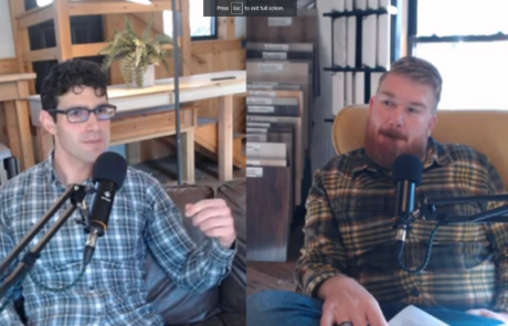 Two men in plaid shirts sit on a couch and talk into microphones in a cozy, wood-paneled room with bookshelves in the background.