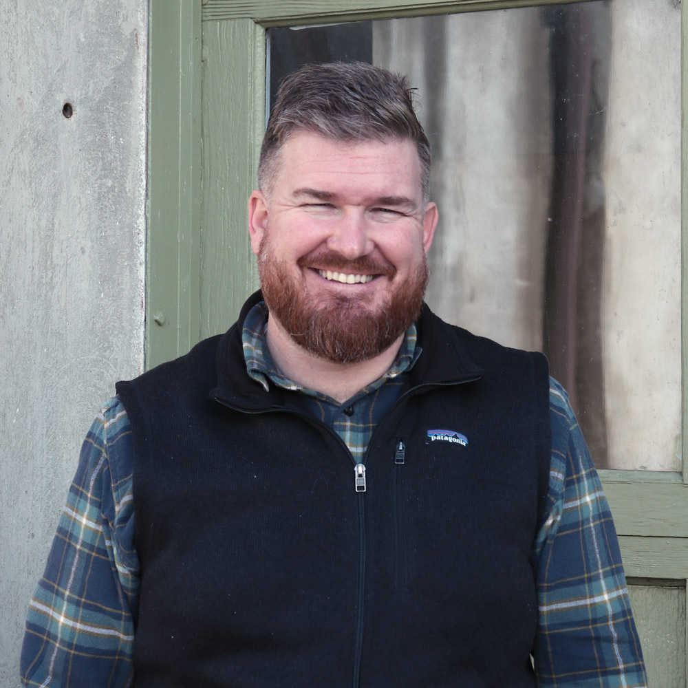 Dave New shot A man with a beard and short hair, wearing a plaid shirt and black vest, stands smiling in front of a green door and window.