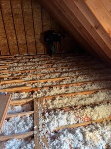 A person works in a wood-paneled attic, spreading insulation material between the joists on the floor, preparing for an upcoming kitchen remodel.