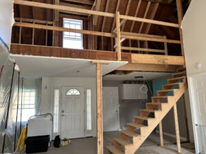 A partially renovated interior with exposed wooden beams, an unfinished loft with stairs, a front door, and a plastic-covered area awaiting a kitchen remodel.