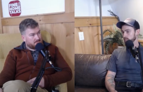 Two men are sitting and talking into microphones during a recording session, one on a couch and the other in an armchair.