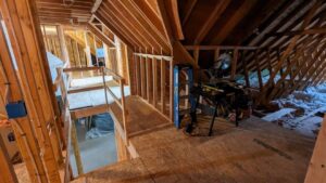 A partially constructed attic with exposed wooden framing, a narrow walkway, and construction tools present on the right side suggests ongoing work, possibly part of a larger kitchen remodel.