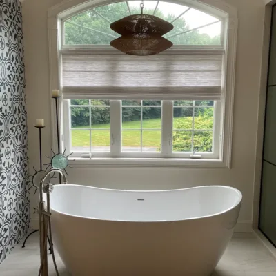 A modern bathroom features a white freestanding bathtub, large window with blinds, patterned accent wall, and a pendant light above.