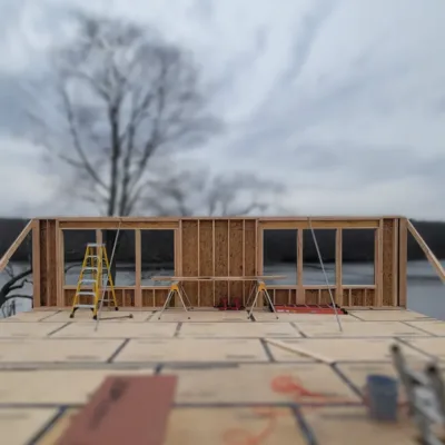 Partially constructed wooden frame of a building with a ladder and tools on the floor, set against a backdrop of a lake and bare trees.