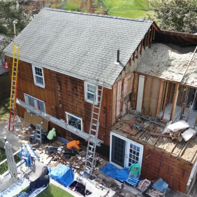 A house under renovation with ladders, debris, insulation, and two workers outside. Part of the roof and walls are removed.