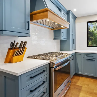 Modern kitchen featuring blue cabinets, white countertops, stainless steel gas stove, knife block, and a window with trees outside.