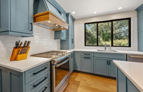 Modern kitchen with blue cabinets, stainless steel appliances, white countertops, wooden flooring, and a large window with a garden view.