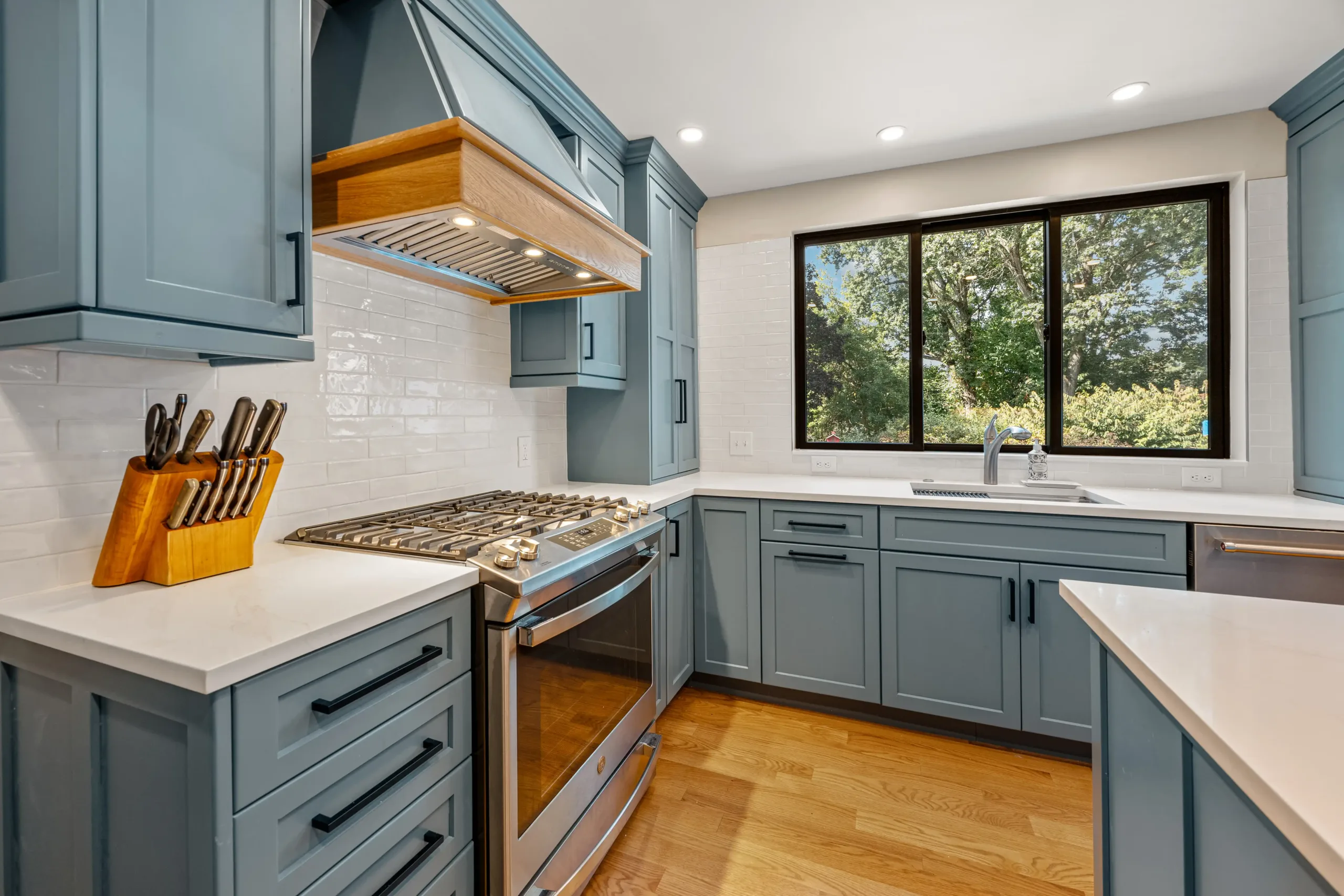 Modern kitchen with blue cabinets, stainless steel appliances, white countertops, wooden flooring, and a large window with a garden view.